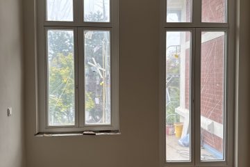 Innenansicht eines renovierten Zimmers mit zwei hohen Rundbogenfenstern, hellen Wänden und einem neuen Parkettboden, teilweise mit Schutzfolie abgedeckt.